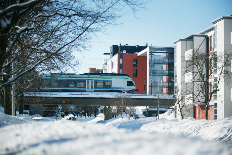 A train travels on a bridge next to buildings in a snowy landscape.