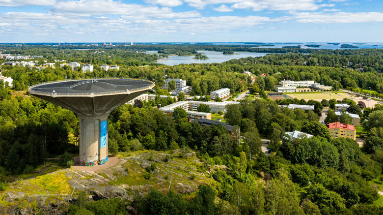 Etualalla on Roihuvuoren vesitorni, jota on kuvattu korkealta ilmasta. Sen takana on kerrostaloja, metsää ja merta.