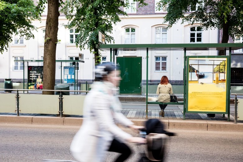 Cyclists travelling along the street so fast that they are slightly out of focus. Behind the bicyclist, a tram stop with a person waiting on the bench.  Trees on both sides of the stop. A row of buildings in the background.